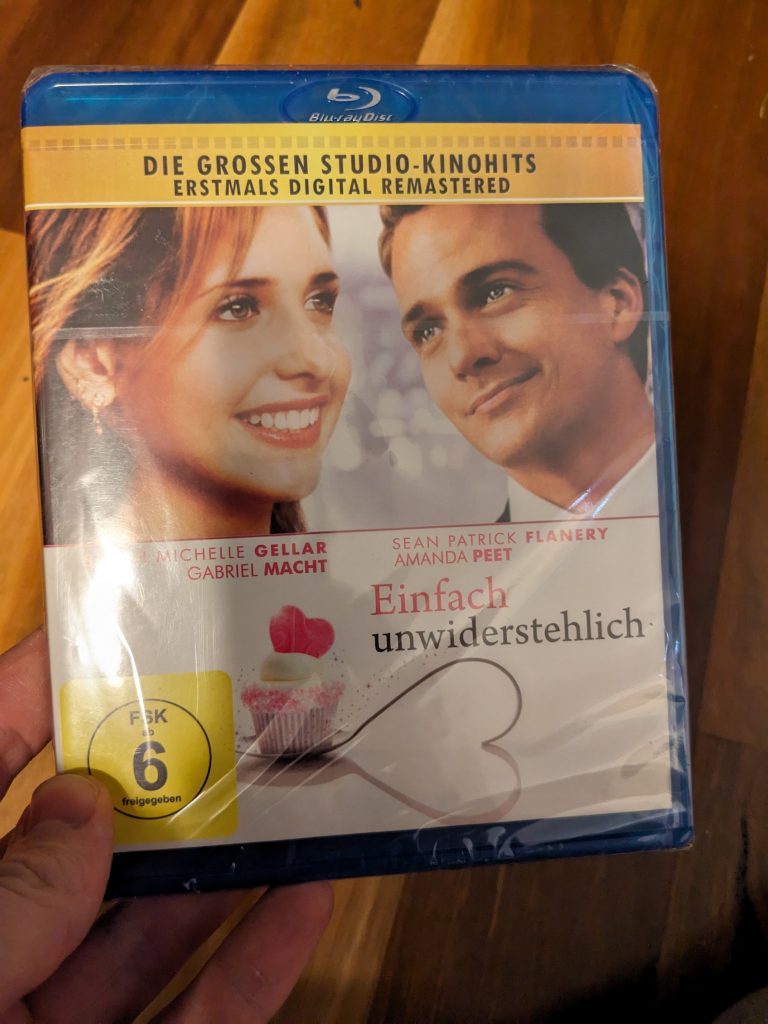 A photo of me holding a shrink-wrapped copy of "Einfach unwiderstehlich", a German-language blu-ray of Simply Irresistable. Sarah Michelle Gellar and Sean Patrick Flannery's heads are on the cover.