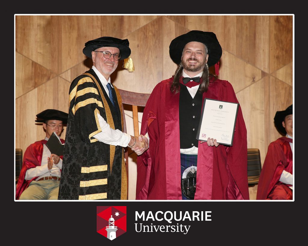 A graduation photo in a Macquarie University frame. I'm wearing crimson doctoral graduation robes and a puffy hat, holding my degree, shaking hands with the Pro-Vice Chancellor Nick Mansfield, who is wearing a fetching gold and black striped number.