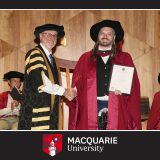 A graduation photo in a Macquarie University frame. I'm wearing crimson doctoral graduation robes and a puffy hat, holding my degree, shaking hands with the Pro-Vice Chancellor Nick Mansfield, who is wearing a fetching gold and black striped number.