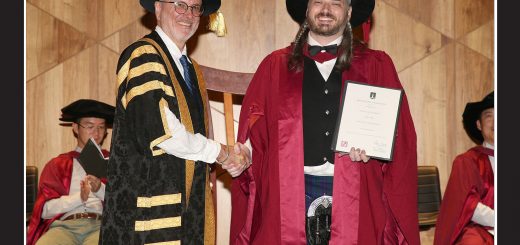 A graduation photo in a Macquarie University frame. I'm wearing crimson doctoral graduation robes and a puffy hat, holding my degree, shaking hands with the Pro-Vice Chancellor Nick Mansfield, who is wearing a fetching gold and black striped number.